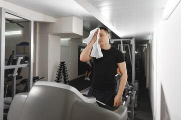 An exhausted man wipes his sweaty face with a hand towel after an intense workout at the gym on the treadmill.