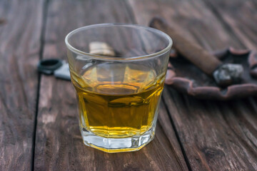 A glass of whiskey stands on a wooden table. Against the background of a cigar with an ashtray out of focus