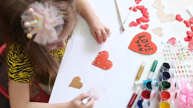 Little girl draws funny hearts with gouache and glitter, Kids art. Crafts concept. How to make a greeting card, Creating handmade gifts for Mothers Day or kids birthday