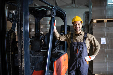 Portrait of experienced young caucasian forklift driver standing by loading cargo machine in storage warehouse.