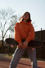 A cute teenage girl in an orange hoodie is posing with a skateboard.