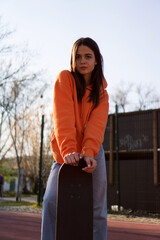 A cute teenage girl in an orange hoodie is posing with a skateboard.