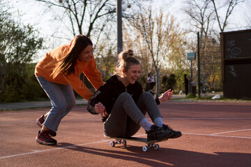 Two young female friends hang out in the city streets. One girl is sitting on the skateboard while the other one is pushing her around.