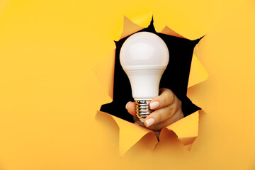 Hand holding an incandescent led light bulb from a torn hole in yellow paper. Concept of saving...