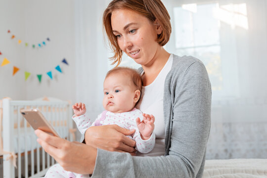 Portrait of a smiling mother holding a baby in her arms and using a smartphone. Childrens room on the background. Concept of teaching and entertaining children using the Internet