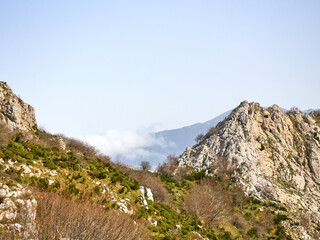 Rocky mountainside with clouds on the ridge and a green forest in spring.