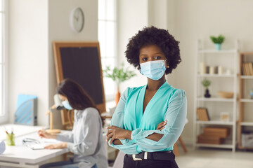 Portrait of black businesswoman with Afro hairstyle in medical face mask standing arms crossed in office. Young woman returns to work after Covid-19 lockdown ends. Coronavirus pandemic safety concept - Powered by Adobe
