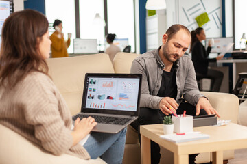 Back view of woman entrepreneur sitting on couch in office using laptop computer while colleagues working on background. Multiethnic coworkers planning new financial project in modern company