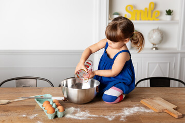 Toddler girl kneeling on kitchen table pouring flower into mixing bowl