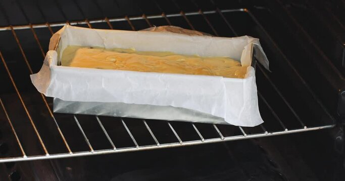 A rectangular baking dish with yellow dough is placed in the oven with one hand