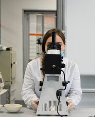 A young woman in a white uniform is working in a laboratory behind a microscope .