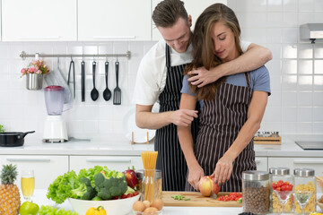 young caucasian man holding his girlfriend while she chop vegetable to prepare lunch in kitchen. couple together concept