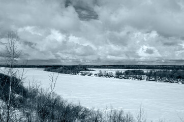 Winter landscape with a high bank of the river covered with ice, cumulus clouds in the sky,.deep snow cover with footprints, a clear horizon line, monochrome, blue tone.