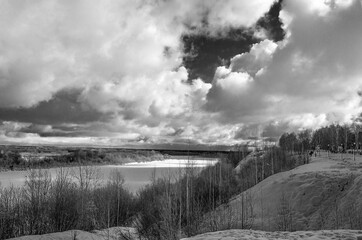 Winter landscape with a high bank of the river covered with ice, cumulus clouds in the sky,.deep snow cover with footprints, a clear horizon line, monochrome.