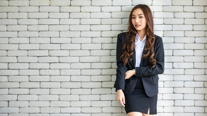 Positive Asian businesswoman wearing classy clothes standing against brick wall in workplace and looking at camera.