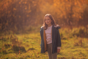 A 10-year-old girl walks in the park in a green coat in autumn.Yellow leaves, a beautiful girl on a walk. Long hair, a smile. Warm orange background.