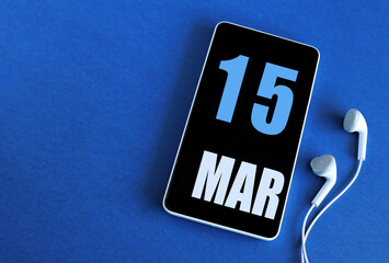 March 15. 15 st day of the month, calendar date. Smartphone and white headphones on a blue background. Place for your text. Springtime month, day of the year concept