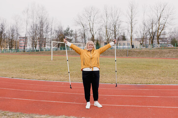 An elderly woman in a yellow sports jacket does physical exercises with Nordic sticks in her hands at the stadium.