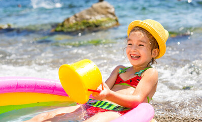Girl in yellow straw hat plays with the wind, water and a water dispenser in an inflatable pool on the beach. Indelible products to protect children's skin from the sun, sunburn. resort at the sea.