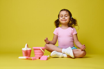 Adorable little girl meditating with closed eyes, sitting near set of beach toys on yellow background with copy space