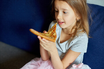 Cute little girl sitting on couch and eating piece of italian Pizza at home