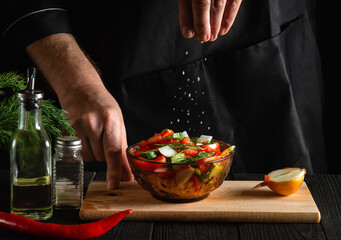 The chef sprinkles salted fresh vegetable salad in a plate on a wooden table. Cooking healthy food in the kitchen in a restaurant