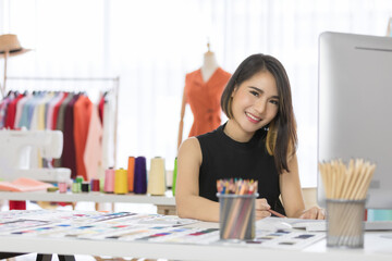 Smiling Asian woman dressmaker is sitting  sketch dress that she has to cut for the customer in the Tailor shop and sewing machines. Concept creating new clothes collection.