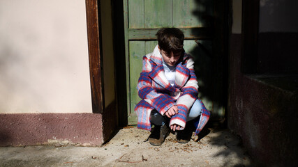 Poor sad small girl with large coat outdoors in front of house, poverty concept.