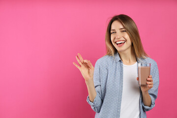 Young woman with glass of chocolate milk showing Ok on pink background. Space for text