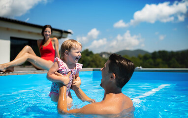 Young family with small daughter in swimming pool outdoors in backyard garden, playing.