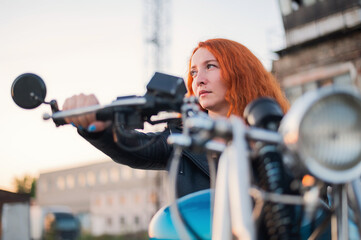 Naklejka na ściany i meble Curly red-haired woman in a black leather jacket sits on a motorcycle. Portrait of a serious girl driving a bike.