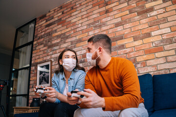 Shooting from below of happy young couple wearing medical protective masks playing video games on console, sitting together on soft sofa in living room. Concept of leisure activity of lovers at home.