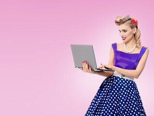 Fototapeta premium Portrait of beautiful woman holding laptop, dressed in pin up style dress in polka dot, over rose pink color background. Blond girl in retro fashion and vintage concept studio image.