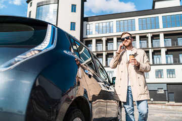 Handsome man with cup of coffee in his hand talking on the phone while standing near his car