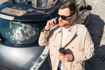 Business man with cup of coffee in his hands talking on the phone while standing near his car