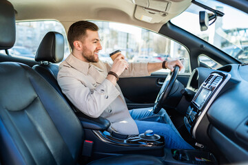 Handsome confident man driving car, holding cup of coffee in hand