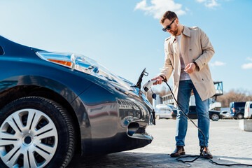 Stylish man in sunglasses inserts the charging cable into the socket of electric car - obrazy, fototapety, plakaty