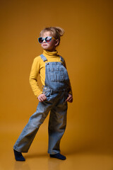 Beautiful little girl wearing stylish denim overall, yellow roll neck jumper and colorful sunglasses standing on bright yellow background. Kids fashion concept
