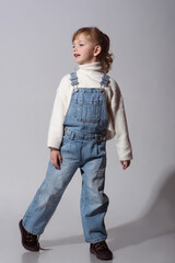 Beautiful little girl wearing stylish denim overall, white roll neck fluffy jumper posing on white background looking back
