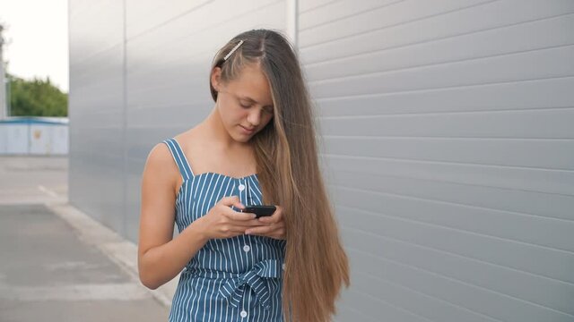 Smiling girl walking down street with mobile phone in her hands. girl looks into phone, types on keyboard text. teenager uses smartphone to communicate. teenager walking down street looks at the phone