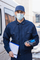 Fototapeta na wymiar arabian courier in medical mask holding payment terminal and clipboard while standing near car