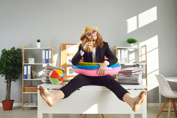 Annual leave and planning travel vacation concept. Funny office worker waiting for summer holidays. Happy barefoot employee in sun hat, sunglasses and swim ring sitting on desk and drinking cocktail - obrazy, fototapety, plakaty