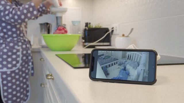 Mother cooking dinner in the kitchen using ip camera as baby video monitor on mobile phone, woman watching her sleeping baby in real-time on smartphone screen. High quality 4k footage