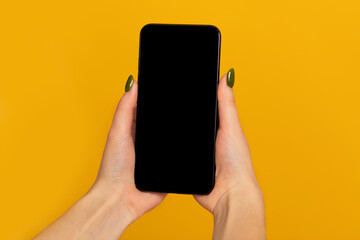 Fototapeta na wymiar Modern smartphone with black blank screen for mockup in woman's hands on yellow studio background