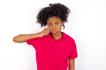 Fototapeta na wymiar Unhappy young beautiful African American woman wearing pink t-shirt against white wall crying while posing at camera whipping tears with hand.