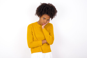 Very bored young beautiful African American woman wearing yellow sweater against white wall holding hand on cheek while support it with another crossed hand, looking tired and sick.