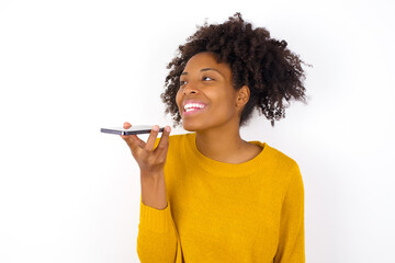 Smiling young beautiful African American woman wearing yellow sweater against white wall sending voice message on her smart phone. Communication and new technologies concept.