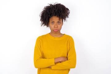 Gloomy dissatisfied young beautiful African American woman wearing yellow sweater against white wall looks with miserable expression at camera from under forehead, makes unhappy grimace