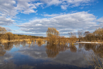 Spring flood, the river overflowed its banks. Sunny spring day. High water level in the river. Rural landscape in early spring. Clouds and trees are reflected in the water.