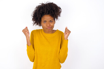 Irritated young beautiful African American woman wearing yellow sweater against white wall blows cheeks with anger and raises clenched fists expresses rage and aggressive emotions. Furious model